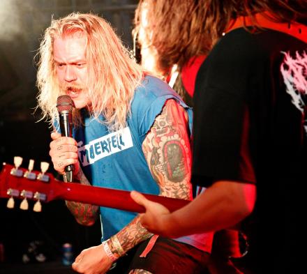 Mand med langt lyst hår, der holder en mikrofon og en mand med den guiar, som står med ryggen til