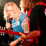 Mand med langt lyst hår, der holder en mikrofon og en mand med den guiar, som står med ryggen til